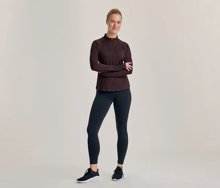Une femme souriante se tient les bras croisés, vêtue d'un haut foncé à manches longues et demi-zip, d'un legging foncé et de chaussures de sport noires et blanches.