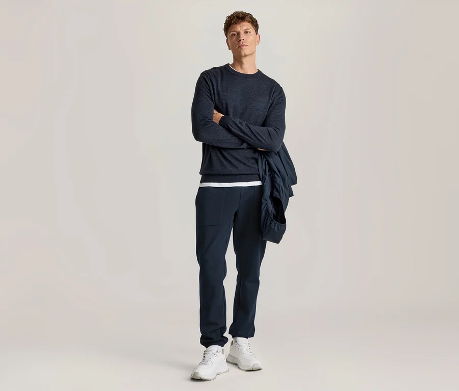 Homme en t-shirt à manches longues bleu foncé, pantalon bleu foncé et baskets blanches, avec une veste bleu foncé drapée sur le bras, les bras croisés.