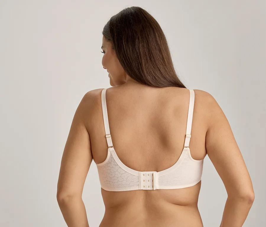 Femme vue de dos, portant un soutien-gorge beige clair à motifs avec une fermeture à trois agrafes. Ses longs cheveux foncés tombent sur ses épaules tandis qu'elle regarde sur le côté.
