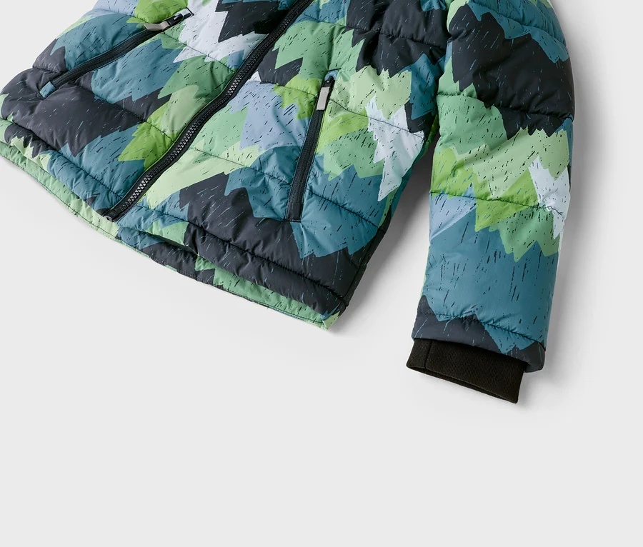 Gros plan d'une doudoune matelassée avec un motif géométrique de formes dentelées vertes, bleues, grises et blanches. La veste a une fermeture éclair noire sur le devant, une poche zippée et un poignet côtelé foncé sur la manche.