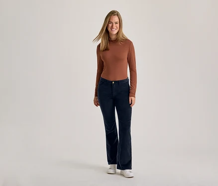 Une femme souriante debout, vêtue d'un haut marron à manches longues, d'un pantalon en velours côtelé bleu foncé et de baskets blanches.