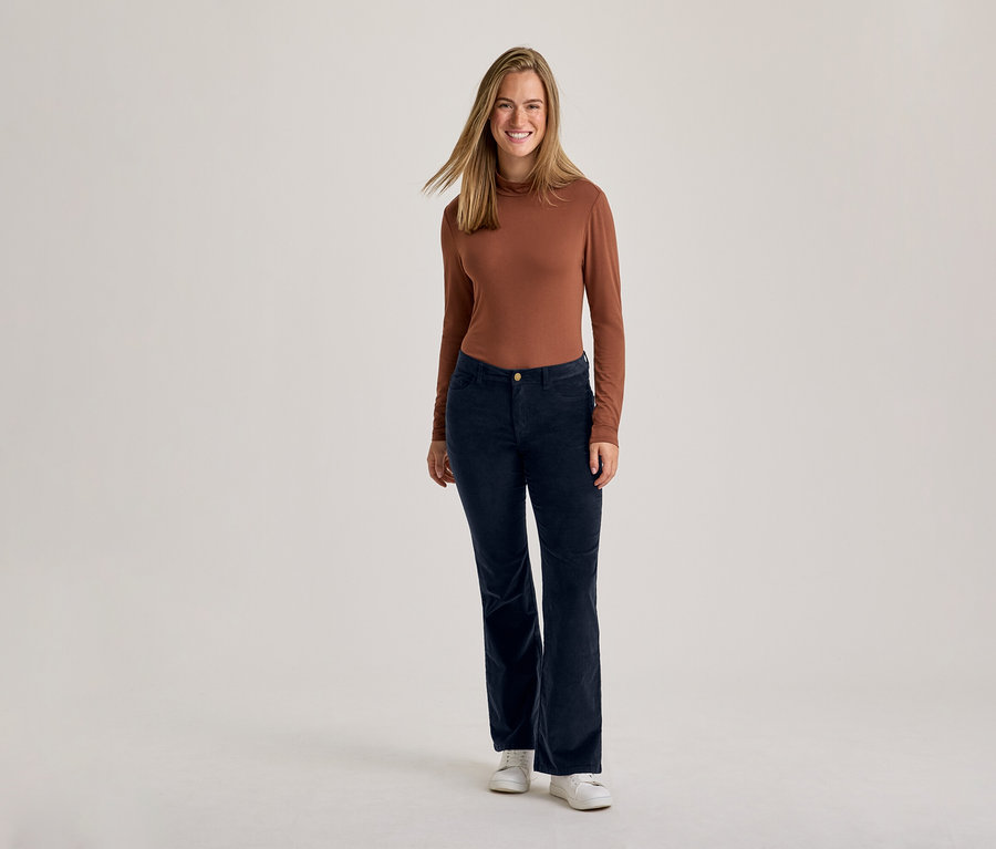 Une femme souriante debout, vêtue d'un haut marron à manches longues, d'un pantalon en velours côtelé bleu foncé et de baskets blanches.