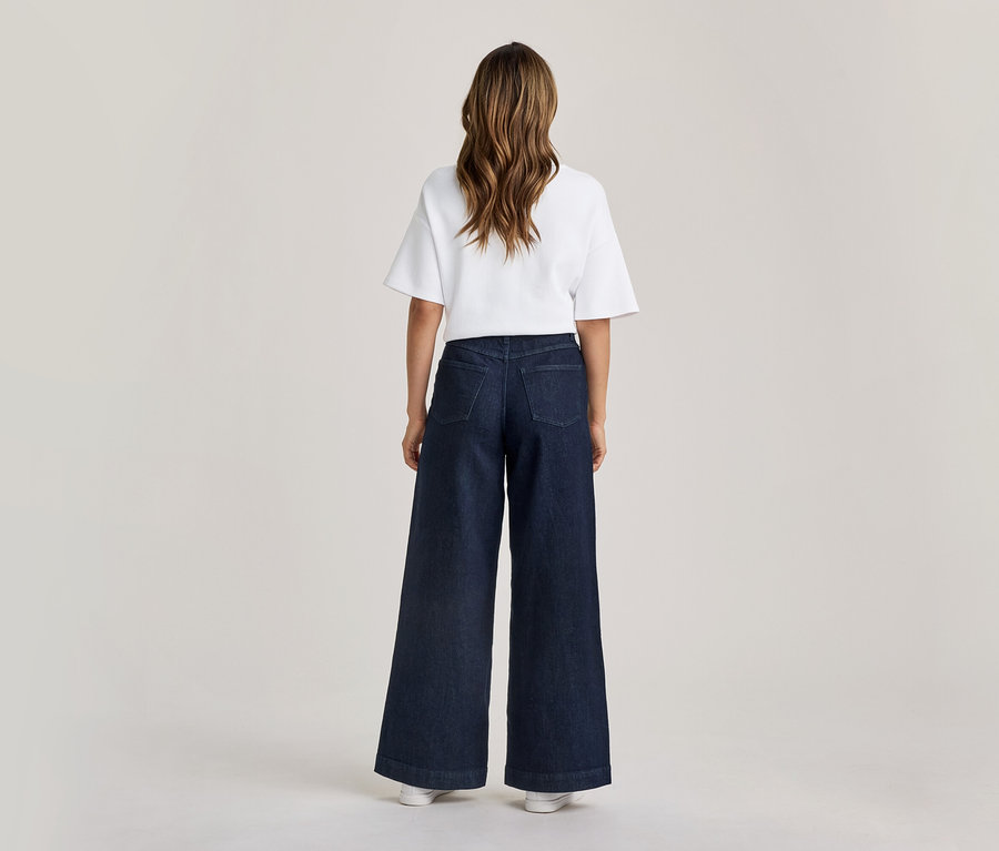 Une personne est debout, dos au spectateur, vêtue d'un t-shirt blanc à manches courtes et d'un jean large bleu foncé. Ses longs cheveux bruns tombent dans son dos.