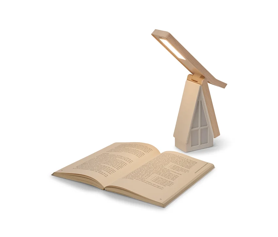 Un livre ouvert est posé devant une lampe de table en forme de maison avec une base beige et un toit en bois. La lampe est allumée, sa lumière LED éclairant le livre.