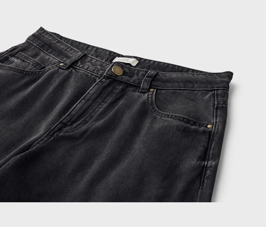 Gros plan sur la partie supérieure d'un jean en denim noir délavé. Le jean a une ceinture avec des passants, une fermeture boutonnée dorée et des poches avant, dont une petite poche à monnaie. Une étiquette blanche avec « MY STYLE » est visible près du bouton.