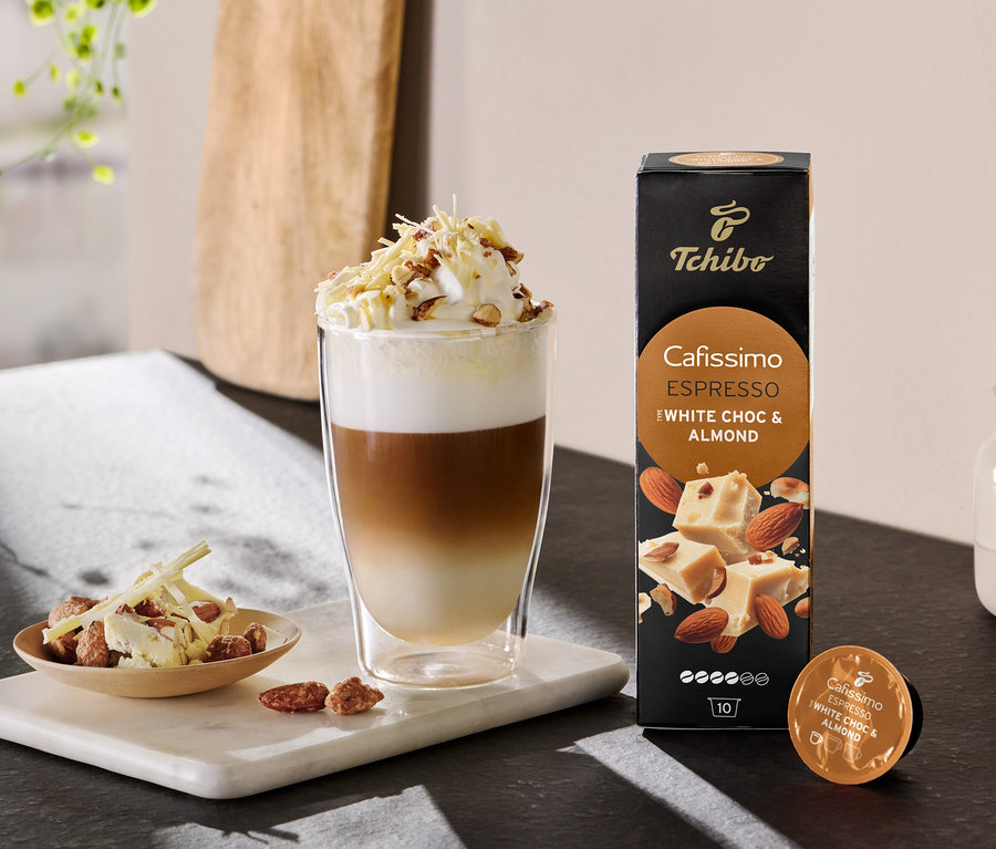 Latte en couches avec de la crème fouettée et des pépites à côté d'un paquet de Cafissimo Flavoured Espresso - White Choc & Almond.