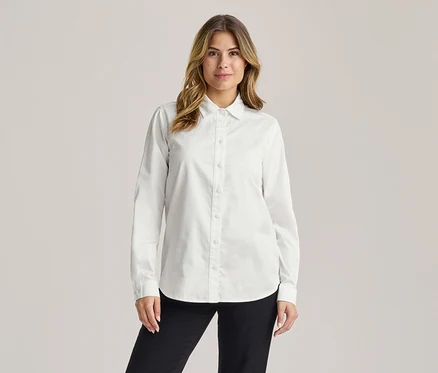 Femme aux longs cheveux bruns ondulés, souriant légèrement, portant une chemise blanche à manches longues et un pantalon noir.