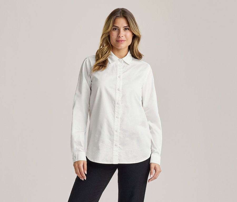 Femme aux longs cheveux bruns ondulés, souriant légèrement, portant une chemise blanche à manches longues et un pantalon noir.