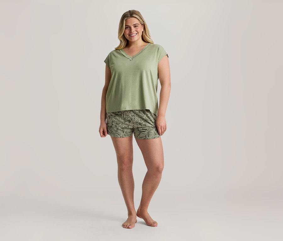 Femme souriante portant un haut vert clair à col en V avec de la dentelle et un short vert olive à motif floral, debout pieds nus.