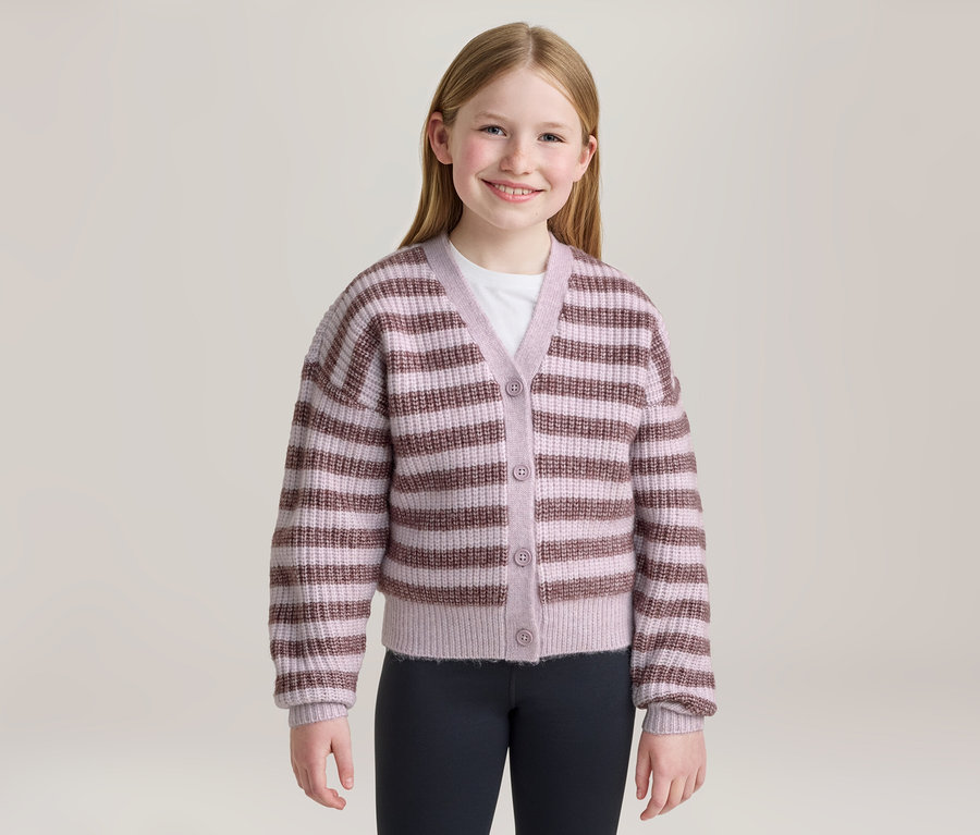 Une jeune fille souriante aux longs cheveux blonds est debout de face, vêtue d'un cardigan rayé rose clair et marron boutonné, d'un t-shirt blanc et d'un legging foncé.