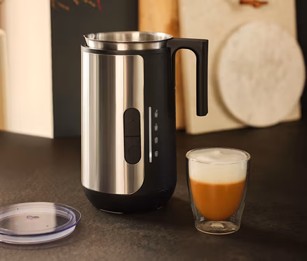 Un mousseur à lait en acier inoxydable est posé à côté d'un verre de café avec de la mousse de lait.