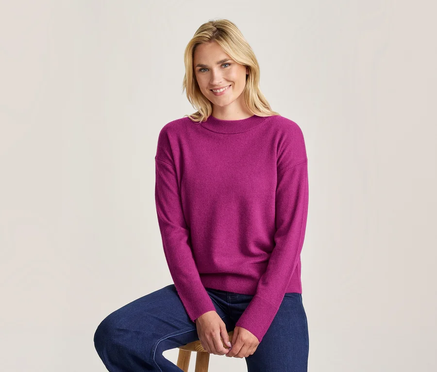 Femme blonde souriante assise sur un tabouret en bois, portant un pull magenta à manches longues et un jean bleu foncé.