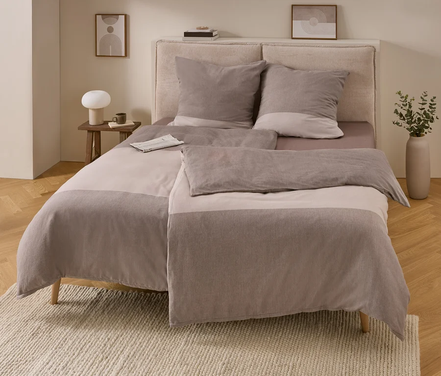 Chambre avec un lit et une parure de lit en flanelle rayée marron clair et taupe. À gauche, une table en bois avec une lampe et une tasse, à droite, un vase avec des plantes vertes. Un journal est posé sur le lit.