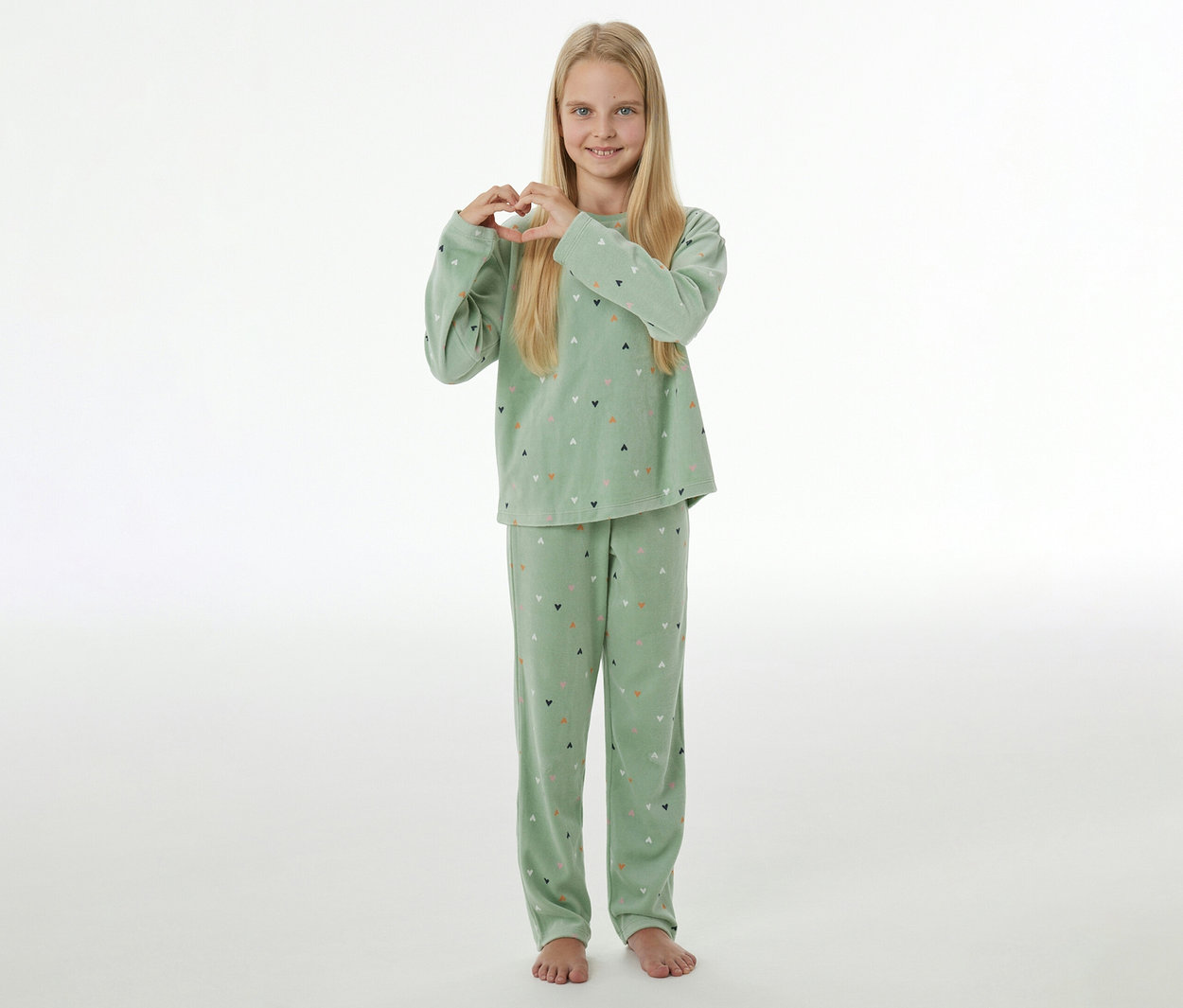 Fille souriante aux cheveux blonds, vêtue d'un pyjama en velours vert clair à motifs de cœurs, formant un cœur avec ses mains.