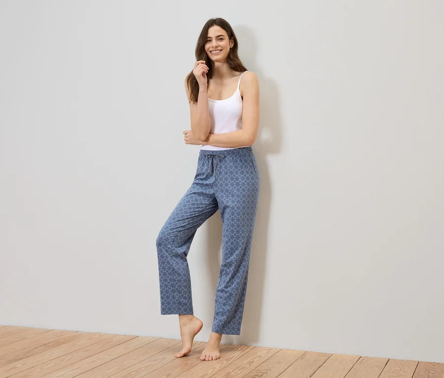 Une femme aux longs cheveux bruns se tient pieds nus devant un mur, portant un débardeur blanc et un pantalon bleu à motifs.