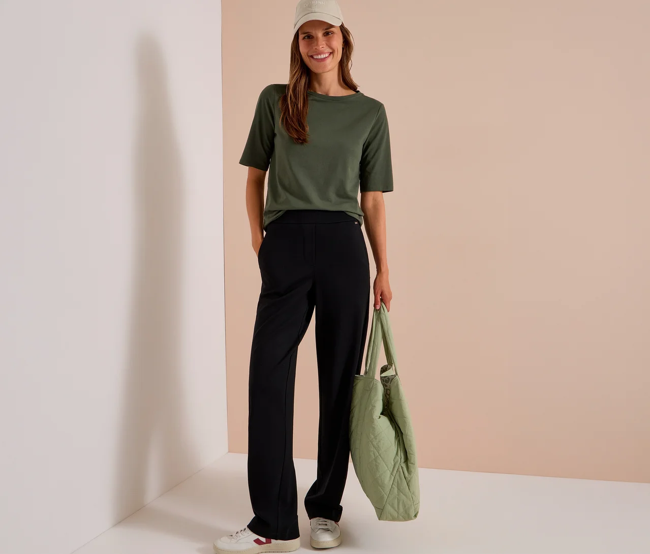 Une femme souriante porte un t-shirt vert olive, un pantalon en tissu CECIL noir, des baskets blanches et une casquette beige, tenant un sac matelassé vert clair.
