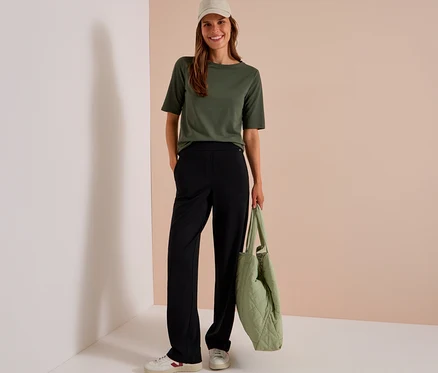 Une femme souriante porte un t-shirt vert olive, un pantalon en tissu CECIL noir, des baskets blanches et une casquette beige, tenant un sac matelassé vert clair.