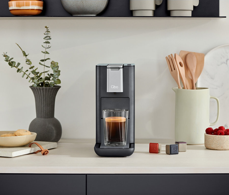 Une machine à café Qbo ESSENTIAL Dark Stone avec une tasse de café. À côté, des capsules et un bol de framboises.