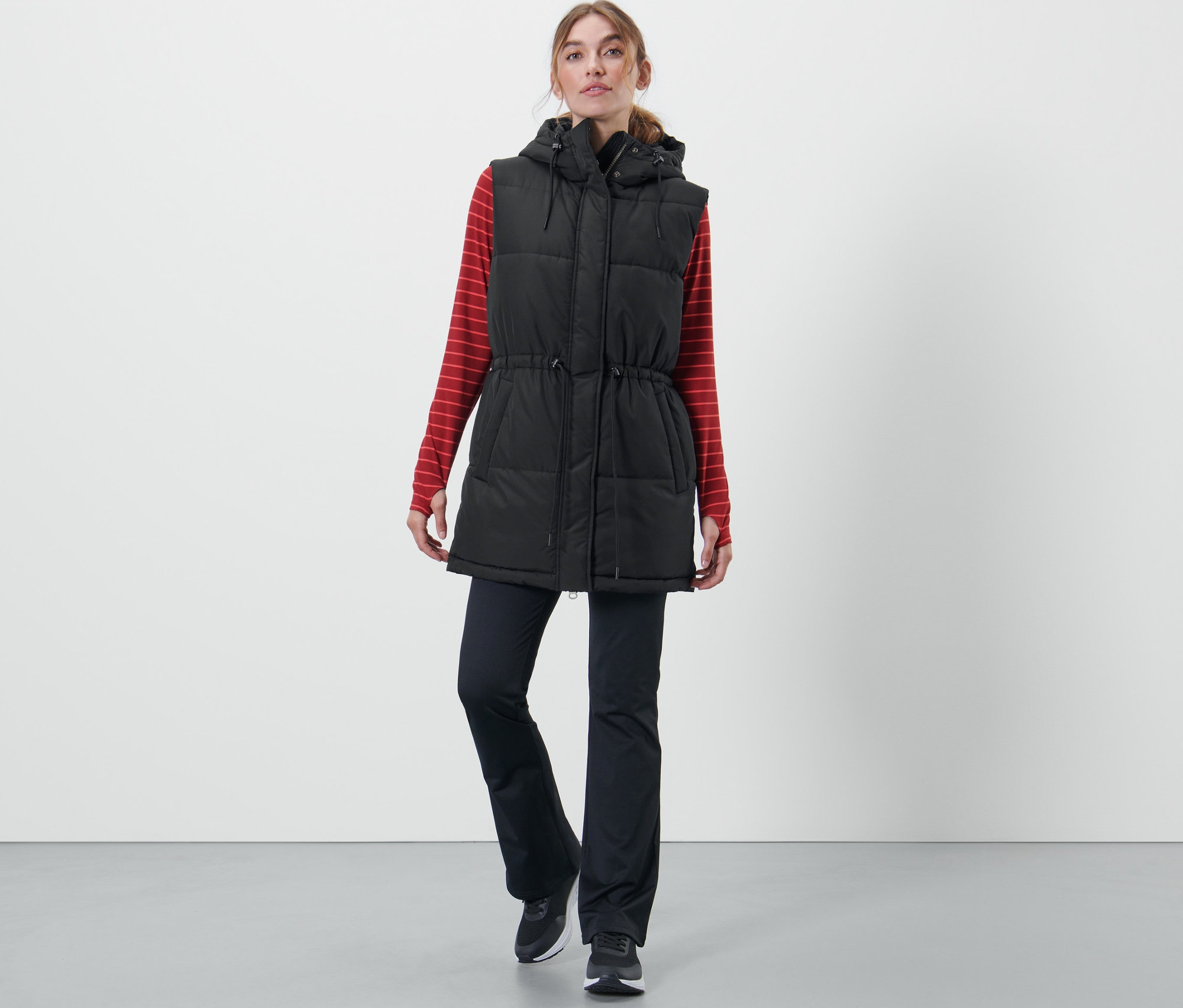 Une femme porte un gilet matelassé à capuche, un t-shirt à manches longues rouge rayé et un pantalon noir.