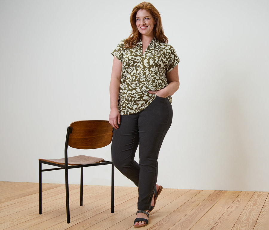 Une femme pose debout, portant une blouse imprimée avec encolure en V et un pantalon en bengaline noir, la main dans la poche, à côté d'une chaise.