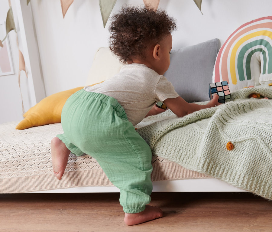 Un bébé grimpe sur un lit et joue avec un Rubik's Cube. Il porte un pantalon à enfiler pour bébé.