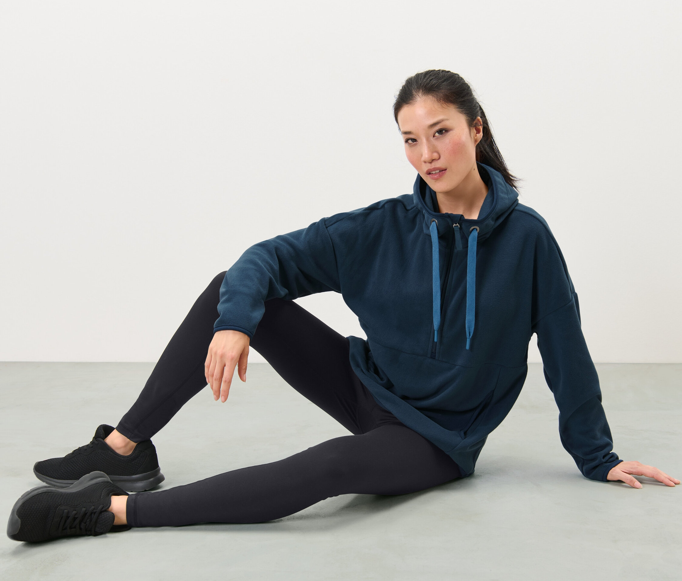 Femme assise par terre portant un pull en polaire à capuche, un legging noir et des baskets.