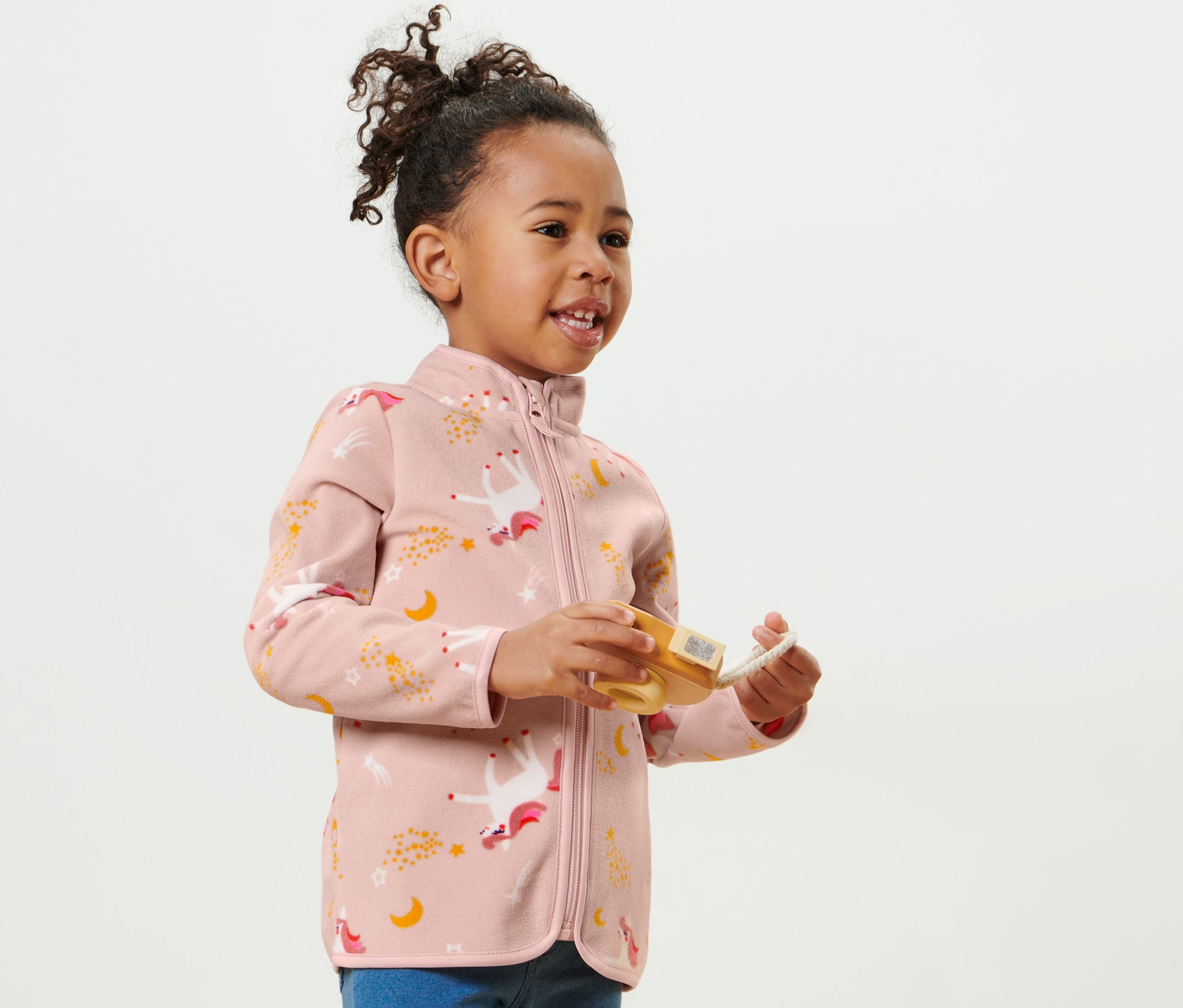 Une fillette souriante porte une jaquette en polaire pour enfant à motif licorne et joue avec un jouet.