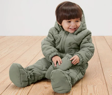 Un enfant souriant assis sur un plancher en bois portant une combinaison d’hiver pour bébé vert olive.