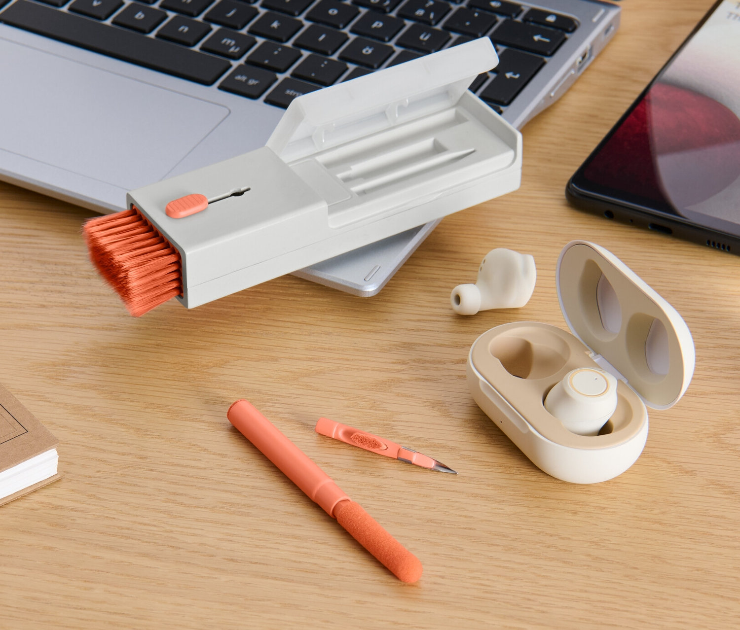 Sur un bureau, on trouve un ordinateur portable, un téléphone, des écouteurs dans un étui, un stylo, un bloc-notes et un set de nettoyage de bureau 4 en 1.