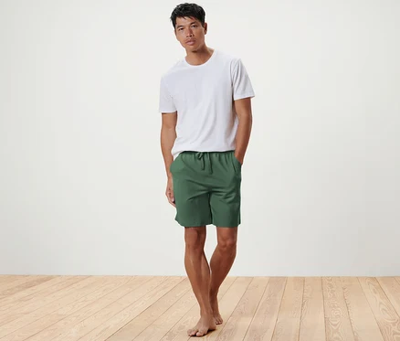 Un homme pose sur un plancher en bois, portant un short de détente olive et un t-shirt blanc.