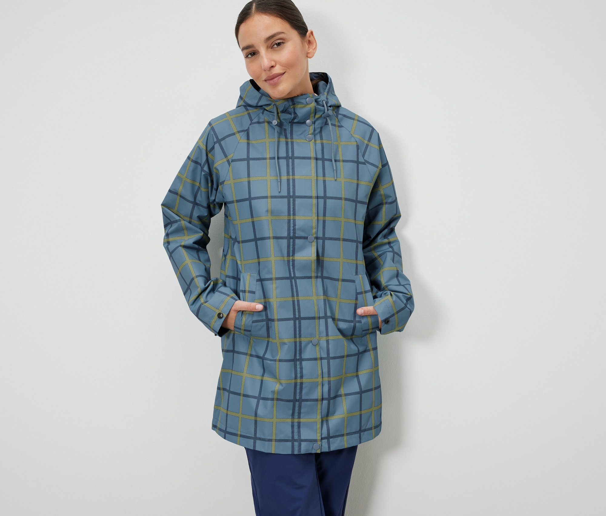 Une femme se tient debout, les mains dans les poches d'une veste de pluie bleue à carreaux.
