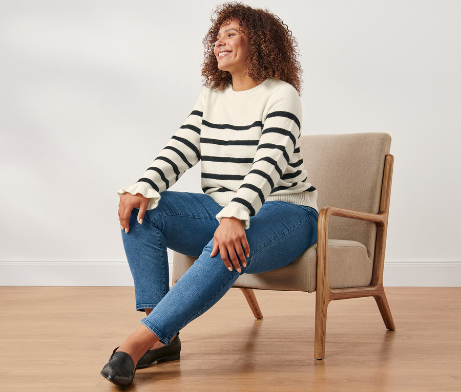 Une femme est assise dans un fauteuil et porte un jean slim et un pull en tricot avec des poignets ruchés à rayures.