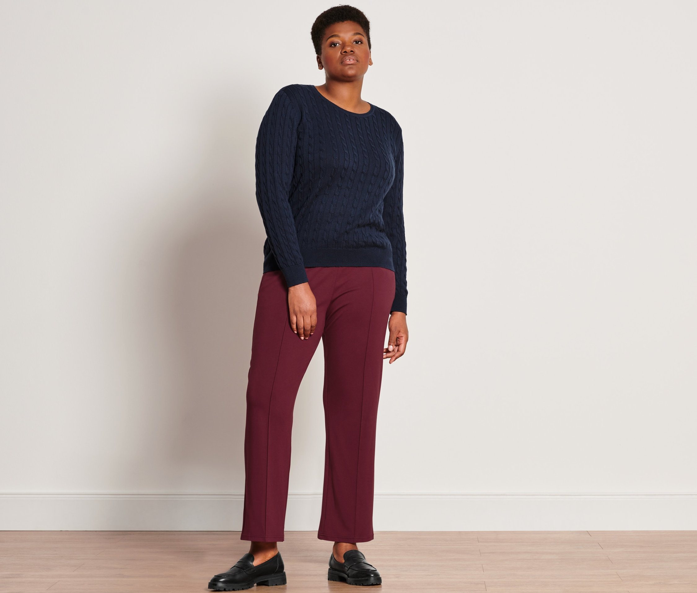 Une femme debout porte un pull en maille torsadée bleu marine, un pantalon de jogging avec nervure et des loafers en cuir.