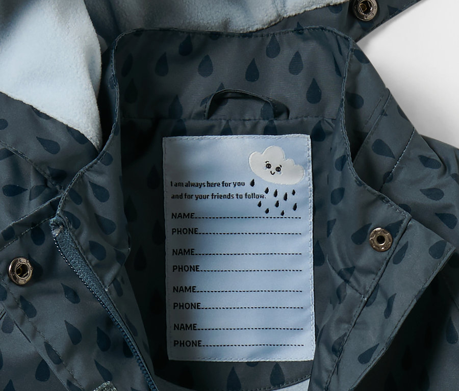 Détail d'une veste de pluie thermique pour tout petit, bleu ardoise à gouttes, avec une étiquette pour le nom et le numéro de téléphone.