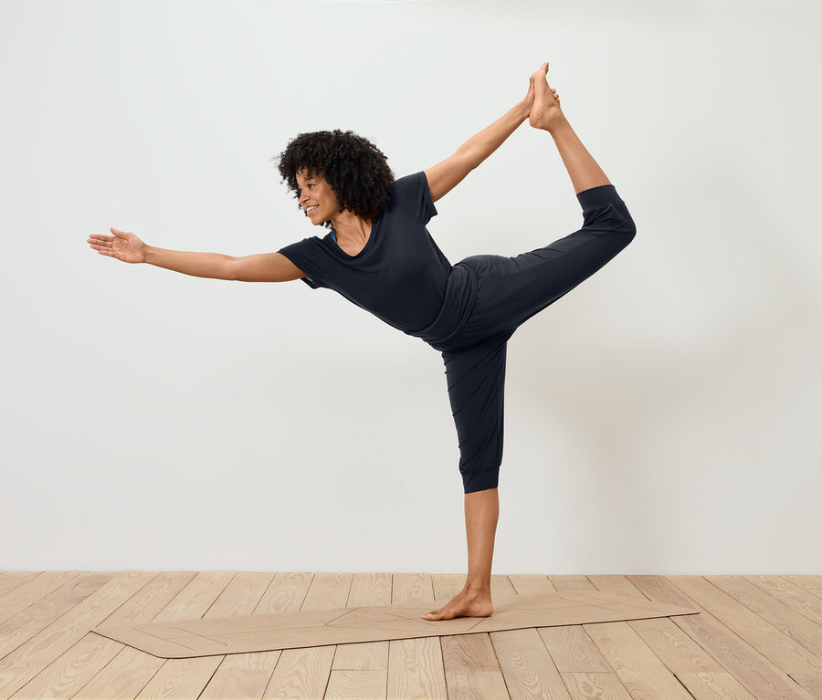 Une femme en tenue de sport noire effectue la pose du danseur sur un tapis de yoga.