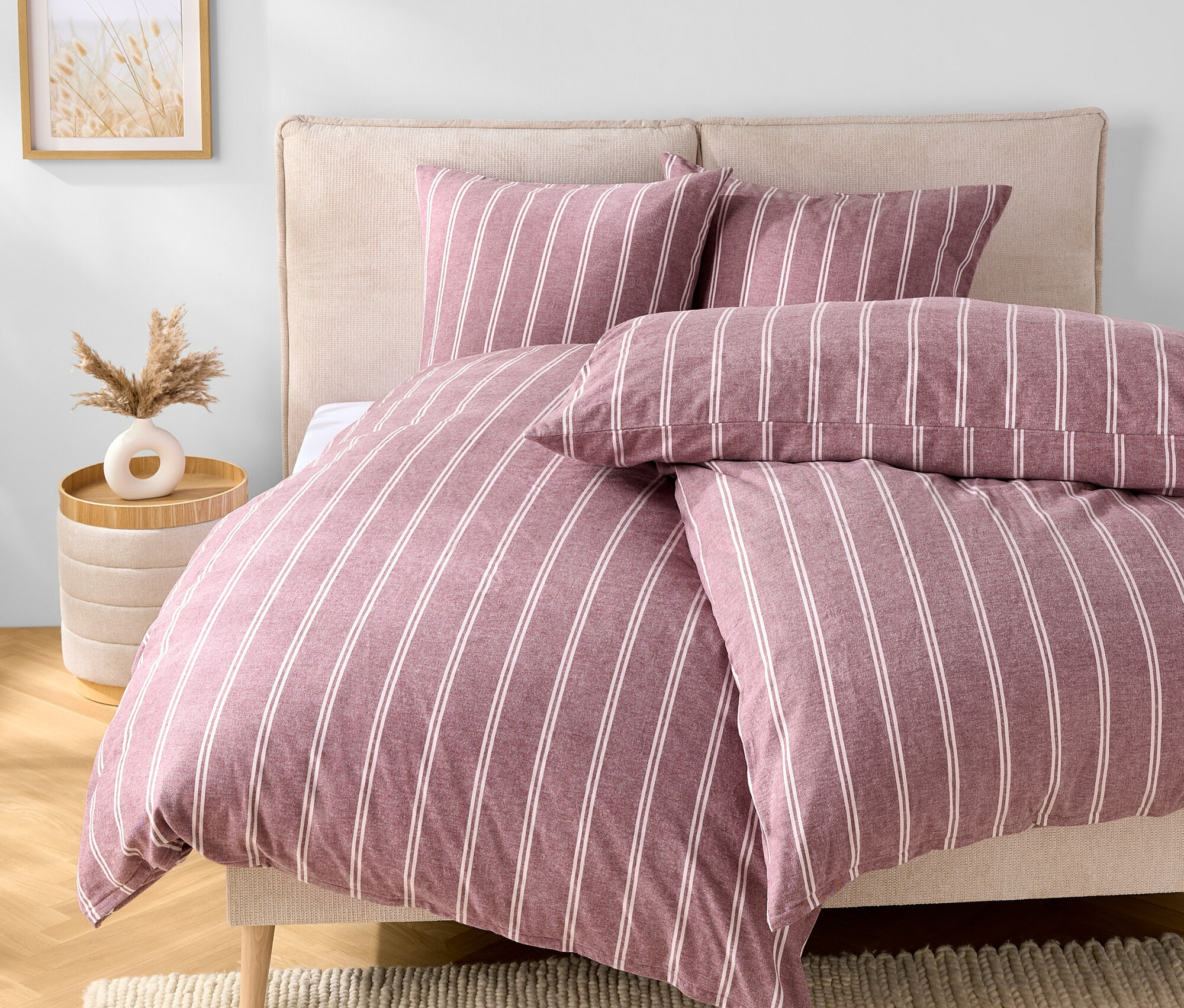 Chambre avec un lit avec des draps rayés violet et blanc, deux oreillers et une tête de lit beige. Une table avec un vase est à côté du lit.