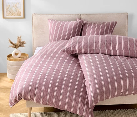 Chambre avec un lit avec des draps rayés violet et blanc, deux oreillers et une tête de lit beige. Une table avec un vase est à côté du lit.