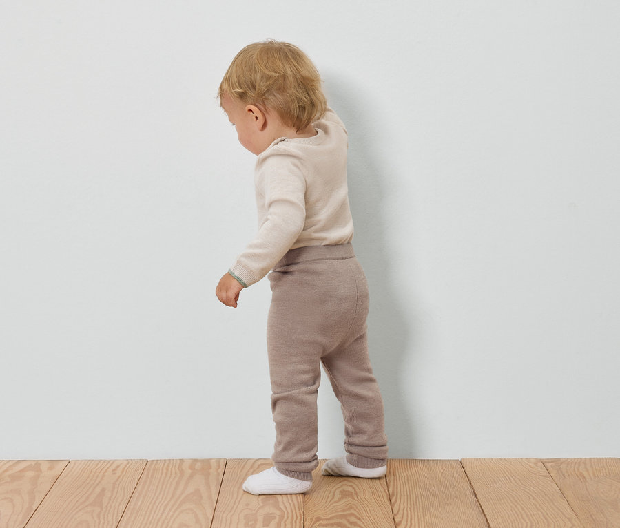 Un bébé portant un pull crème et un pantalon marron se tient dos à un mur blanc.