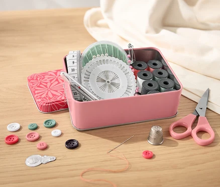 Boîte en métal ouverte contenant un nécessaire à couture, des boutons, des ciseaux et un mètre ruban sur une table en bois.