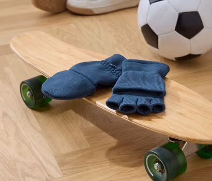 Des gants bleus à doigts reposent sur une planche à roulettes en bois avec des roues vertes. À l'arrière-plan, il y a un ballon de football et des chaussures.