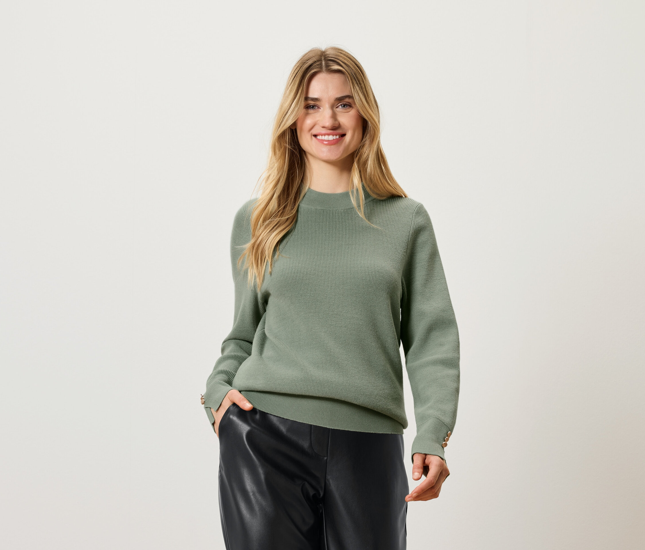 Une femme aux cheveux blonds pose en pull olive et pantalon en cuir noir.