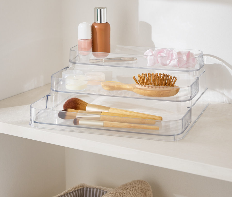 Organisateur de maquillage transparent à trois niveaux sur une étagère blanche à côté de paniers avec des serviettes.