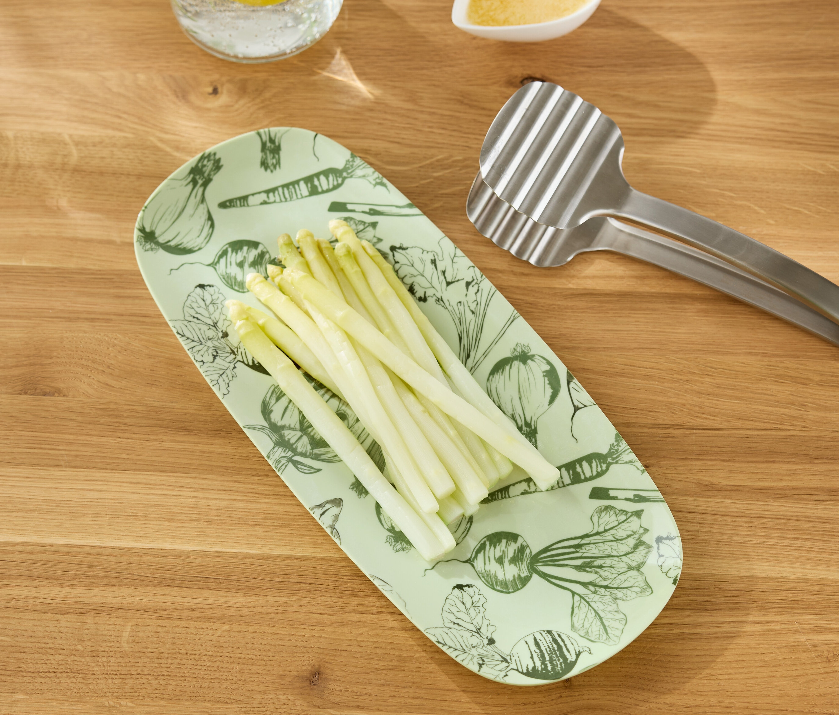 Des asperges blanches sont disposées sur un plat de service allongé vert clair avec un imprimé de légumes, à côté d'une pince à grill argentée sur une table en bois.