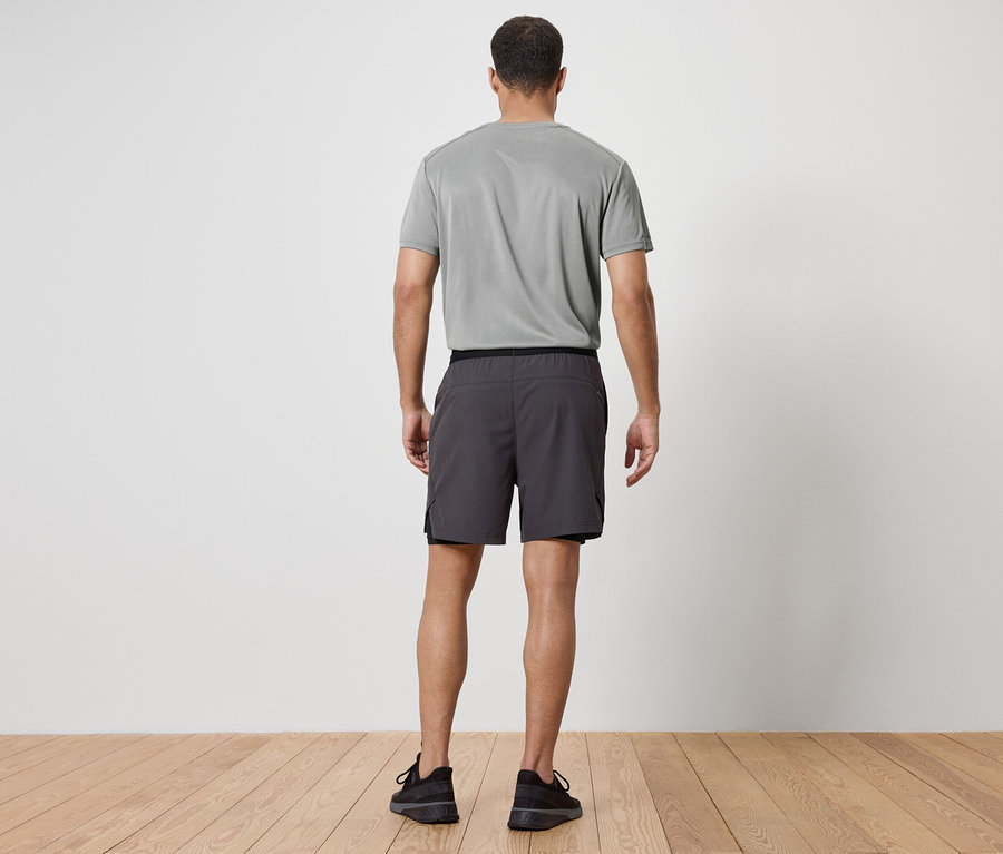 Un homme, dos à la caméra, porte un t-shirt gris, un short gris et des baskets noires.