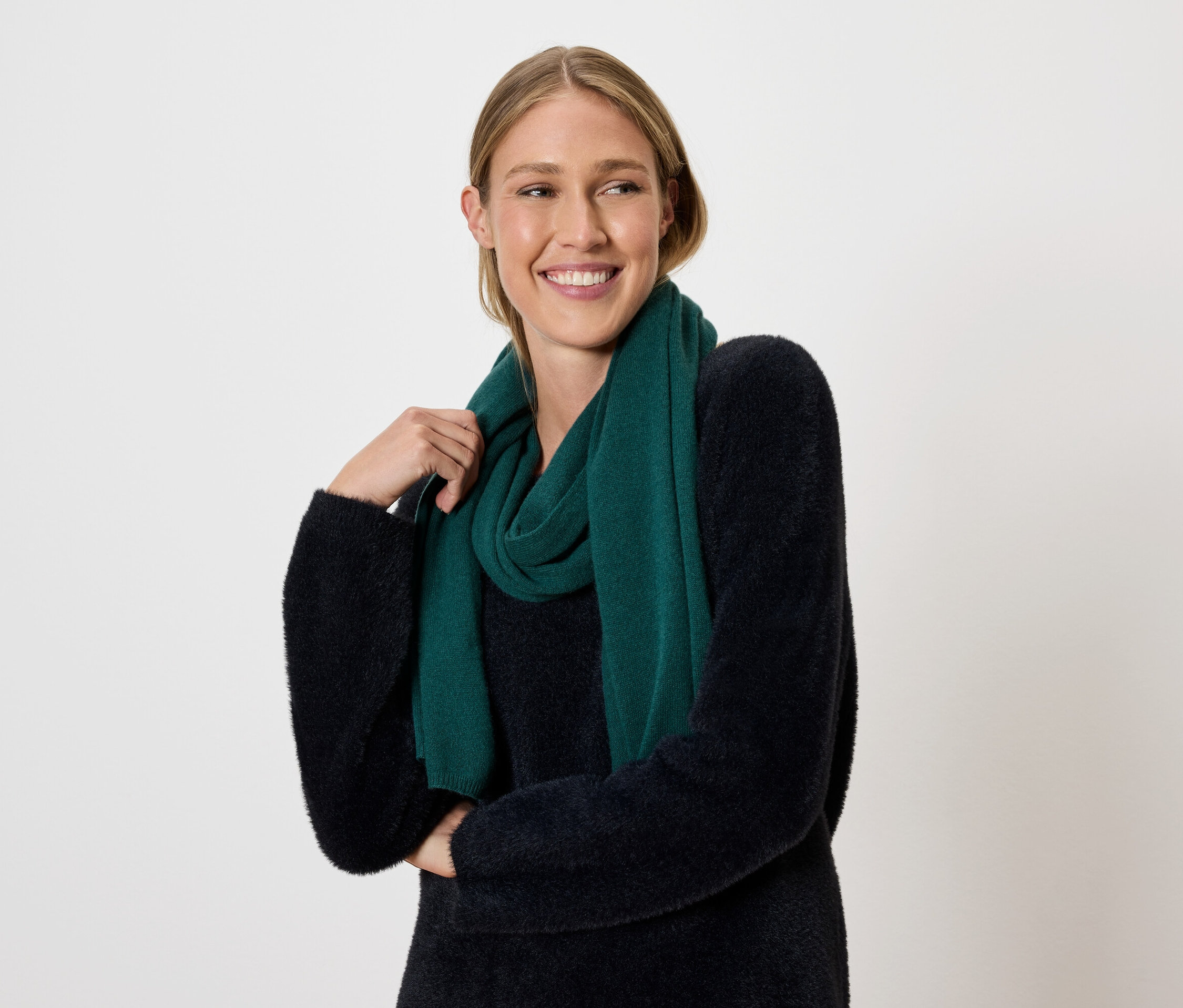 Une femme blonde souriante pose sur un fond clair avec une écharpe vert foncé et un pull.