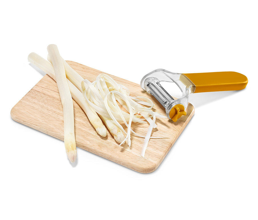 Sur une planche à découper en bois reposent trois tiges d'asperges blanches, des copeaux d'asperges et un éplucheur à légumes jaune et argenté.