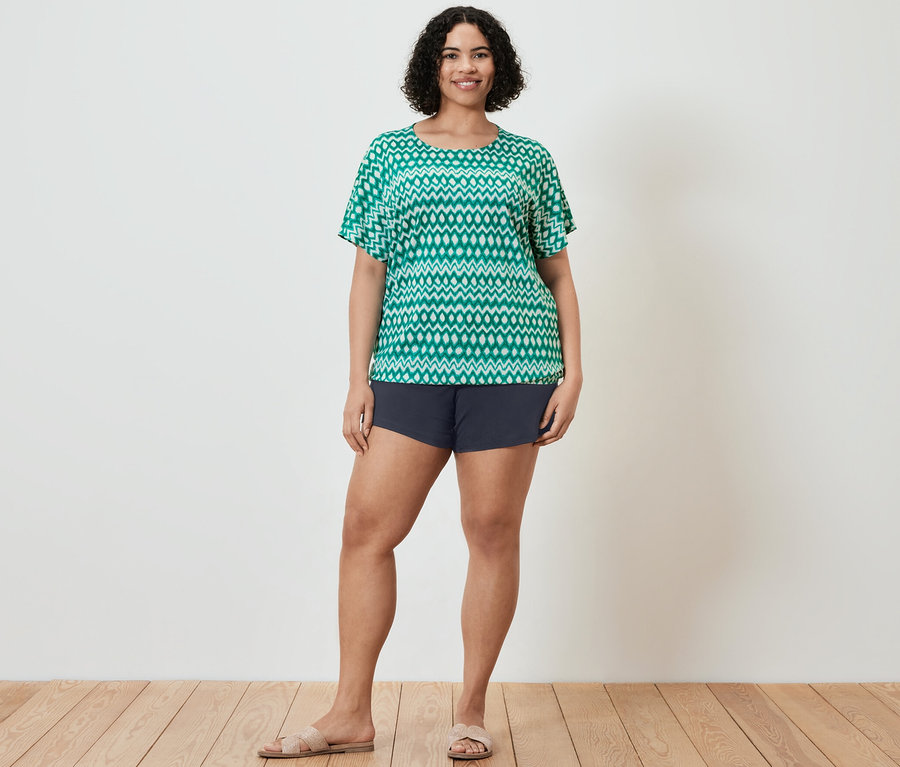 Une femme pose, portant un haut vert imprimé et un short bleu marine. Elle porte aussi des sandales.