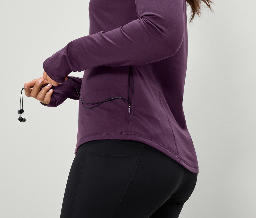 Une femme tient des écouteurs, porte une veste de sport violette et un legging noir.