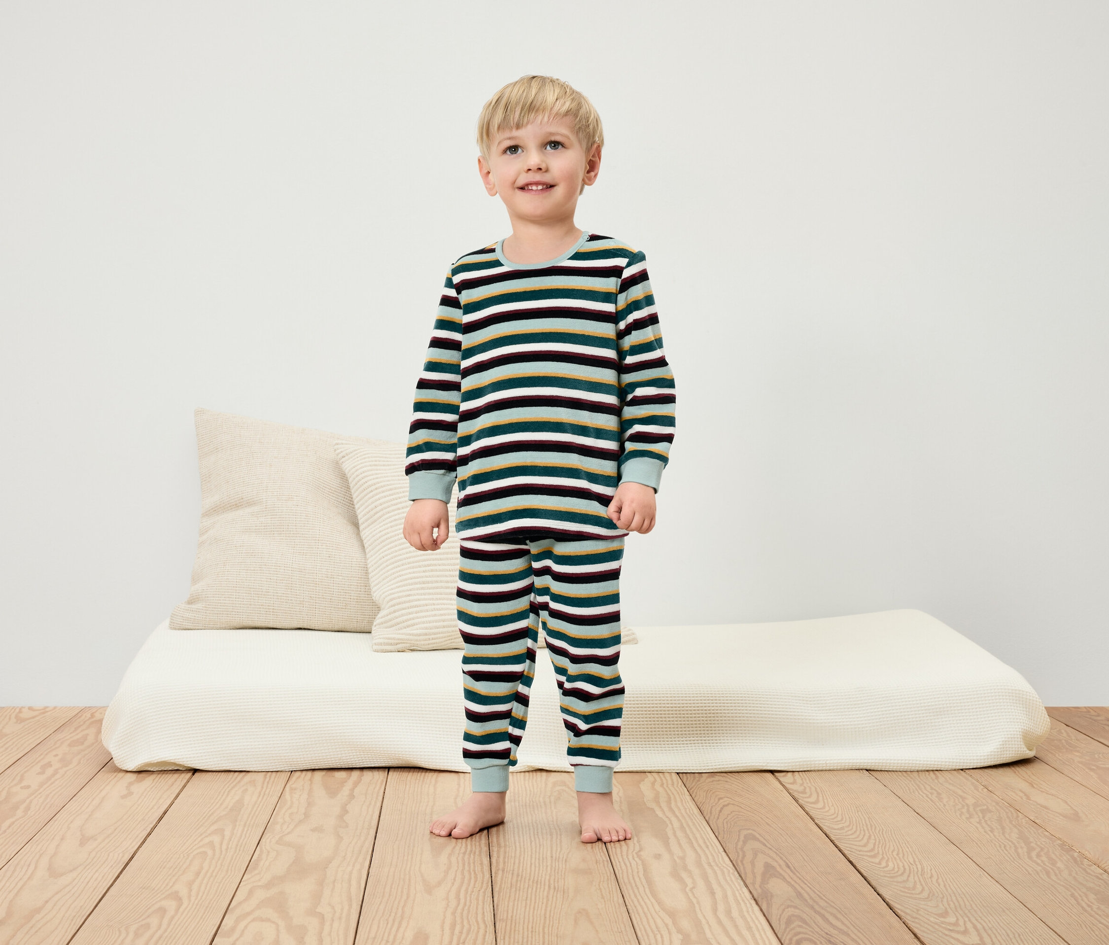 Un garçon en pyjama rayé se tient pieds nus sur un plancher en bois devant un mur blanc.