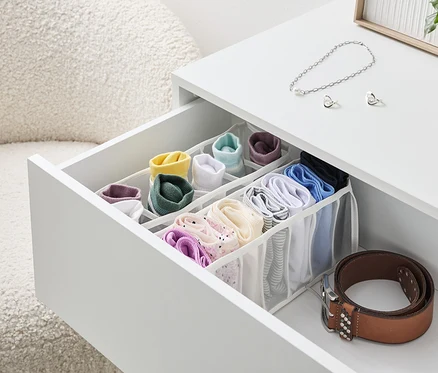 Un tiroir ouvert d'une table blanche avec un organisateur rempli de chaussettes et de sous-vêtements colorés, à côté duquel se trouve une ceinture marron.